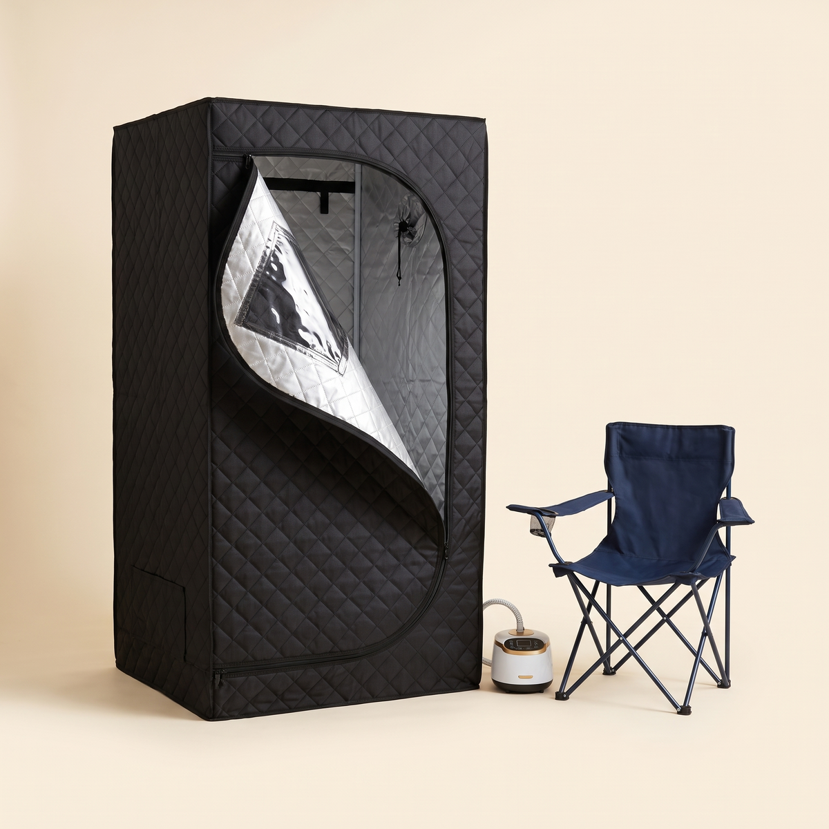 Black quilted portable steam sauna tent with clear window beside a folding chair on a cream studio background